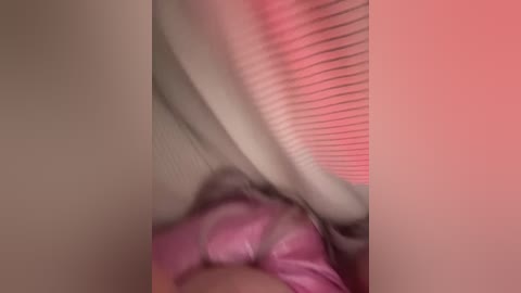 A blurred video shows a pink object, possibly clothing, partially covering a person's body. The background has vertical blinds with a pink hue. The image is slightly out of focus.