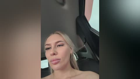 Video of a fair-skinned blonde woman with straight hair, wearing minimal makeup, sitting in the back seat of a car. The background features the car's interior and a blurred window.