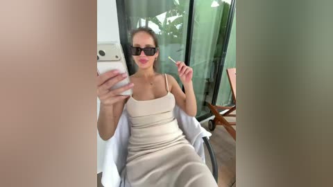Video of a light-skinned woman with straight, dark hair wearing sunglasses and a beige dress, sitting indoors with a white phone in hand, holding a toothpick.