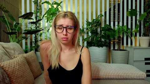 Video of a blonde woman with glasses in a black tank top, surrounded by plants, in a cozy living room with vertical blinds and a patterned sofa.
