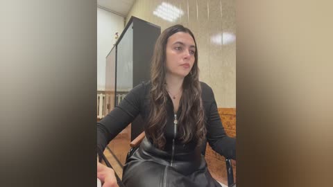 Video of a young woman with long, wavy brown hair, wearing a black leather jacket, seated in a wheelchair, indoors with beige wallpaper, wooden floor, and a glass door in the background.