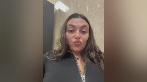 A video of a young woman with long, wavy brown hair, making a pouty face. She wears a black zip-up jacket and is standing in a beige-tiled bathroom with a mirror and fluorescent lighting.