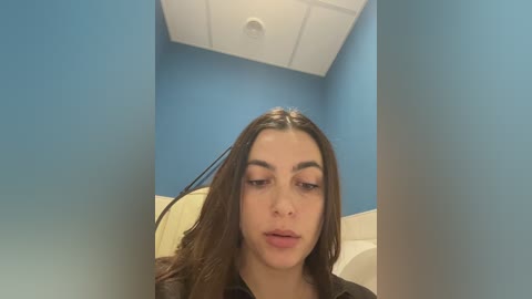 Video of a young woman with long brown hair, light skin, and full lips, wearing a black jacket, seated in a blue-walled room with white ceiling and a beige chair.