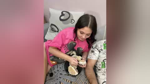 Video of a woman with long dark hair, wearing a pink t-shirt, lying on a bed with a gray and white patterned blanket, holding a pair of shoes.