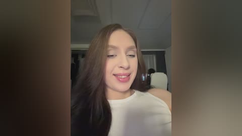 Video of a smiling, fair-skinned woman with long, dark brown hair, wearing a white sleeveless top, standing indoors near a doorway. Background shows blurred people and white walls.