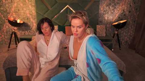 A video depicts two women in robes in a retro bedroom with tiled walls, green bed, and lamps. One is blonde, the other brunette, both with neutral expressions.