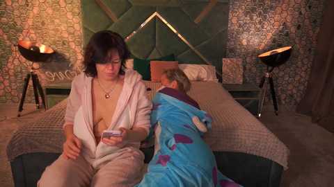 Video of a woman in a pink robe reading a book on a bed, while a child in a blue nightgown sleeps beside her in a cozy bedroom with patterned wallpaper and modern lamps.