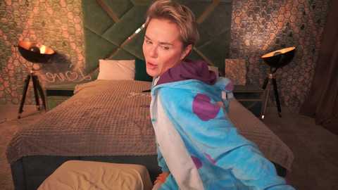 Video of a woman in a blue, purple, and white bear onesie standing beside a beige bed with green, patterned wallpaper.