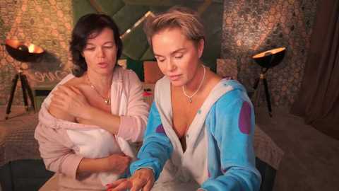 Video of two women in a cozy room with dim lighting. One with short dark hair in a light pink shirt, the other with short blonde hair in a blue hoodie, both leaning over a table.