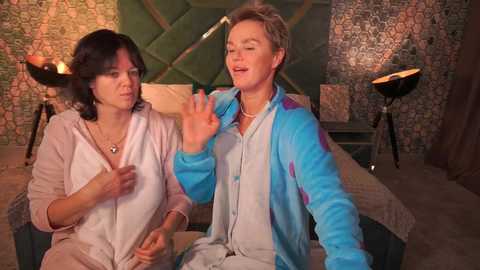 Video of a middle-aged woman with short dark hair in a light pink robe, sitting next to a man in a blue and white jacket, both smiling and waving, in a dimly lit room with green and brown patterned wallpaper and two black floor lamps.