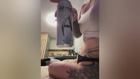 Video of a woman with fair skin, wearing a white tank top and black panties, holding a spray can, sitting on a carpeted floor, with visible tattoos on her thighs and lower abdomen, in a cluttered room with wooden furniture.