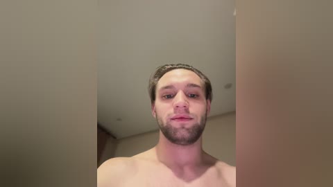 Video of a shirtless, young Caucasian man with short brown hair, a trimmed beard, and light skin, standing indoors with a neutral expression.