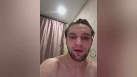 A video of a topless, bearded white man with light skin and short brown hair, looking directly at the camera with a neutral expression. Background includes a pink curtain and a small circular mirror.