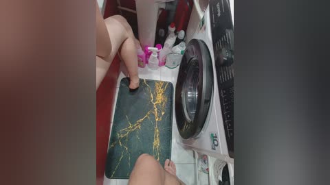 A video from an overhead perspective shows a nude woman standing on a green, marble-patterned mat in a laundry room. The room features various cleaning products and laundry items scattered around.