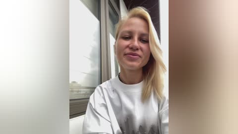 Video of a blonde woman with fair skin and straight hair, wearing a white shirt, standing indoors near a glass window, with blurred faces in the background.