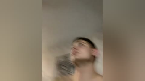 A blurry video of a young man with short dark hair, seen from the side, looking upward. He appears shirtless, with a blurred background of beige walls.