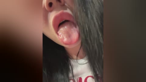 A close-up video of a woman's partially open mouth, with her tongue extended and slightly wet. She has long, dark hair and is wearing a white T-shirt with red text. The background is blurred.