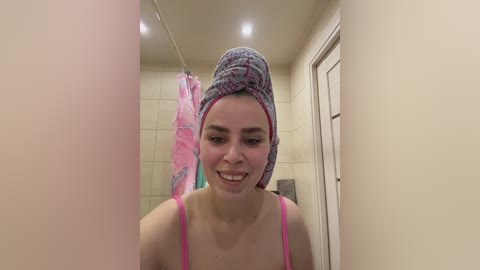 Video of a young woman with fair skin and brown hair, wrapped in a pink towel, standing in a tiled shower with a pink shower curtain.