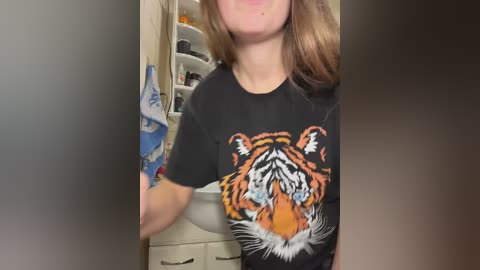 A video of a woman with light skin and wavy brown hair, wearing a black t-shirt with a tiger graphic, standing in a kitchen with open cabinets in the background.