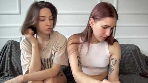 Video of two young women with fair skin, one with shoulder-length brown hair, the other with straight, light brown hair. They sit side by side on a gray couch, wearing casual white tops. Background features a white paneled wall.