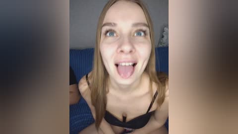 A video of a young Caucasian woman with fair skin, long blonde hair, and blue eyes, wearing a black bra, sticking out her tongue and smiling. She is indoors, on a blue couch, with a blurred background.