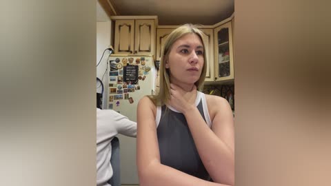 Video of a young blonde woman with fair skin, wearing a sleeveless gray top, in a cluttered kitchen with wooden cabinets and a refrigerator covered in magnets.