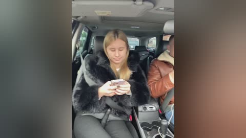 Video of a young woman with blonde hair, wearing a black faux fur coat, black pants, and a white top, seated in a car, texting on a phone, with another person in the background.