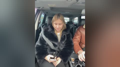 Video of a blonde woman in a black faux fur coat, sitting in a car, holding a smartphone, next to a man in a brown leather jacket.