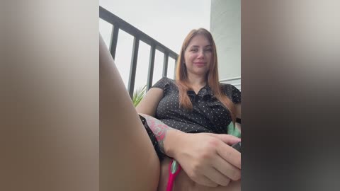 A video of a young woman with fair skin, straight red hair, and a medium build, wearing a black polka-dotted blouse, seated outdoors with legs spread, revealing a red and pink thong.