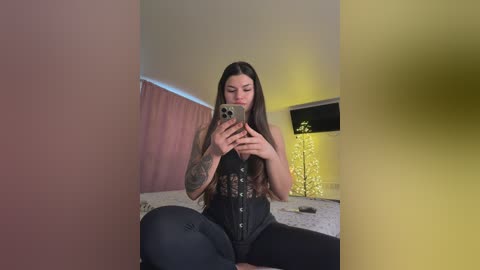 Video of a tattooed woman with long hair, wearing a black corset and tight jeans, taking a selfie in a dimly lit room with a yellow lamp and pink curtains.