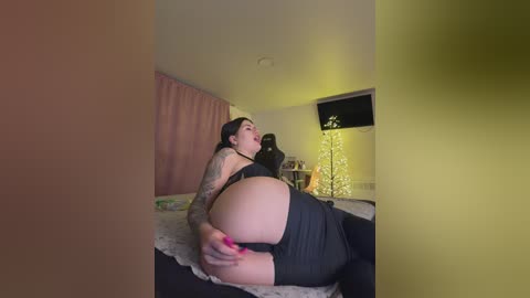 A video of a curvy, tattooed woman with dark hair and pink nails, wearing a black dress, lying on a bed in a dimly-lit, cozy room with a lit Christmas tree.