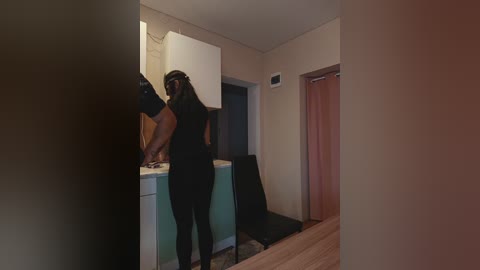 A video of a woman in a black dress and mask, leaning on a kitchen counter, in a dimly lit room with beige walls, wooden floor, and a white refrigerator.