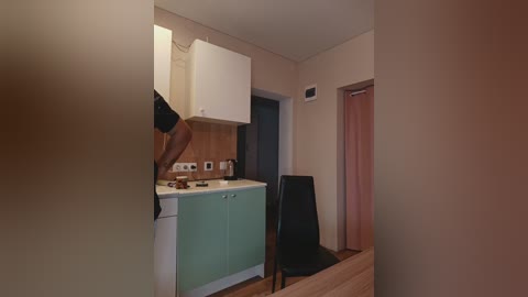 A video of a compact, beige-walled kitchen with a white countertop, a small green cabinet, a black chair, and a partially visible person in a black shirt.
