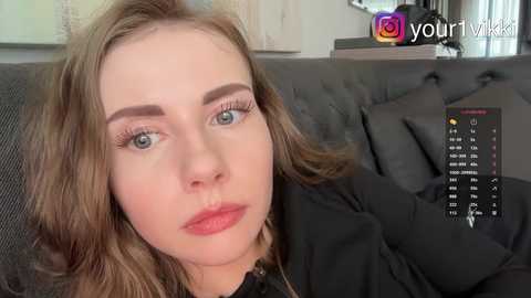 A video of a fair-skinned woman with light brown hair, blue eyes, and a neutral expression, wearing a black top, sitting on a dark grey couch. The background shows a TV screen with a colorful social media icon and a small, colorful decorative object.