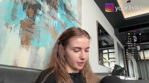 Video of a young woman with long, light brown hair, wearing a black dress, sitting on a grey couch in a modern, minimalist room with abstract blue and gold artwork on the wall.