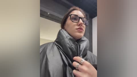 A video of a woman with glasses, fair skin, and brown hair, wearing a black puffy jacket, standing indoors against a beige wall with a dark ceiling.