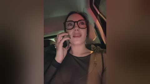Video of a woman with dark hair, wearing black-rimmed glasses and a black top, holding a phone, seated in a car with dim lighting.