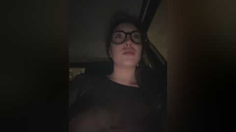 Video of a woman with glasses, wearing a black top, lying on a dark surface, likely inside a car.