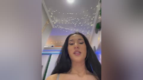 Video of a young woman with long black hair, wearing a grey top, standing in a modern, dimly lit room with white walls and starry ceiling.