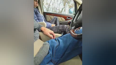 Video of an older man in a car, wearing a blue puffer jacket and gray pants, resting his hand on the steering wheel.