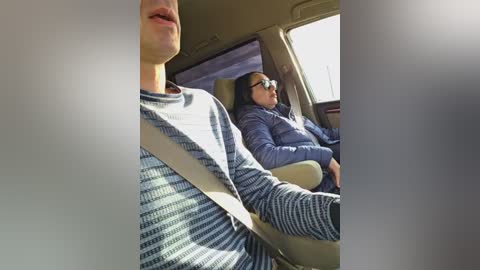 Video of two people in a car; a young man with a striped sweater and a woman with glasses and a jacket, both wearing seatbelts, looking out the window.