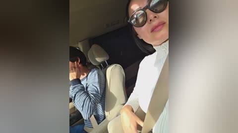 Video of a woman with fair skin, black sunglasses, and a white top, sitting in a beige car seat. A man with dark hair and a striped sweater is visible in the background.