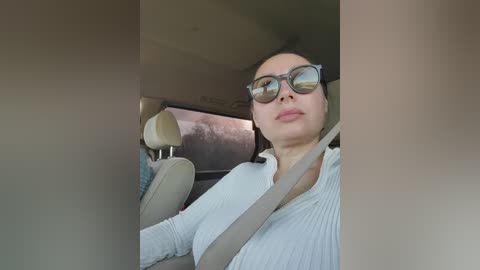 Video of a middle-aged woman with fair skin, wearing large sunglasses, a white blouse, and a seatbelt, driving a beige car with beige seats. The background shows a blurred, sunlit landscape.
