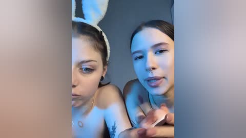 Video of two young women with fair skin and light makeup, one with bunny ears, smiling. They have tattoos and are topless, holding makeup brushes.