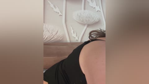 Video of a woman in a black tank top, seen from behind, with her brown hair tied up. She stands in front of white, textured wall art featuring abstract, seashell-like designs.