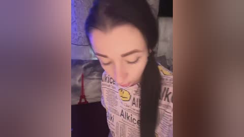 Video of a young woman with fair skin and long black hair, wearing a yellow and white newspaper-patterned dress, looking down. Background includes a dimly lit room with a red candle and a beige wall.