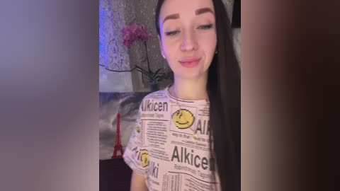 A video of a young woman with long black hair, wearing a white T-shirt with yellow smiley faces and newspaper headlines. She stands against a dimly lit wall with a purple orchid plant.