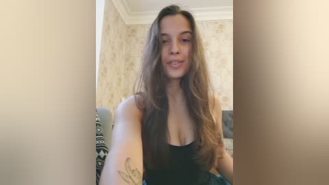 Video of a young, light-skinned woman with long brown hair, wearing a black tank top, sitting on a gray couch with patterned cushions. The background shows beige wallpaper with floral patterns.