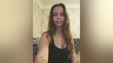 Video of a young woman with long, wavy brown hair, wearing a black tank top, smiling, standing in a room with beige wallpaper and a patterned sofa.