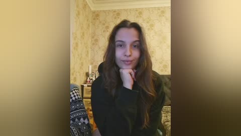 Video of a young woman with long, wavy brown hair, fair skin, and a slight smile, resting her chin on her hand, seated on a patterned couch in a cozy, wallpapered room with a fireplace.